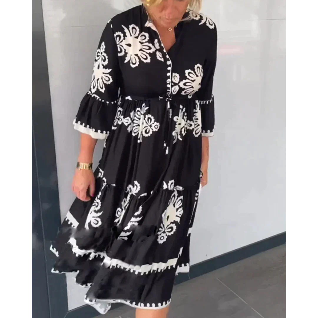 Abito nero con motivi floreali bianchi, maniche a tre quarti e gonna a balze. Donna bionda in posa su sfondo neutro. Moda elegante e casual.