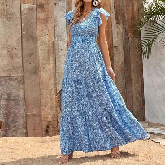 Donna con abito lungo estivo azzurro a fiori, in piedi su sabbia, sfondo di legno e pianta. Moda estiva, abbigliamento femminile elegante.