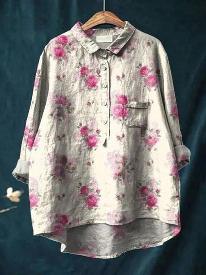 Camicia donna in lino grigio con stampa floreale rosa, maniche lunghe, colletto classico, taschino frontale, appesa su gruccia di legno.