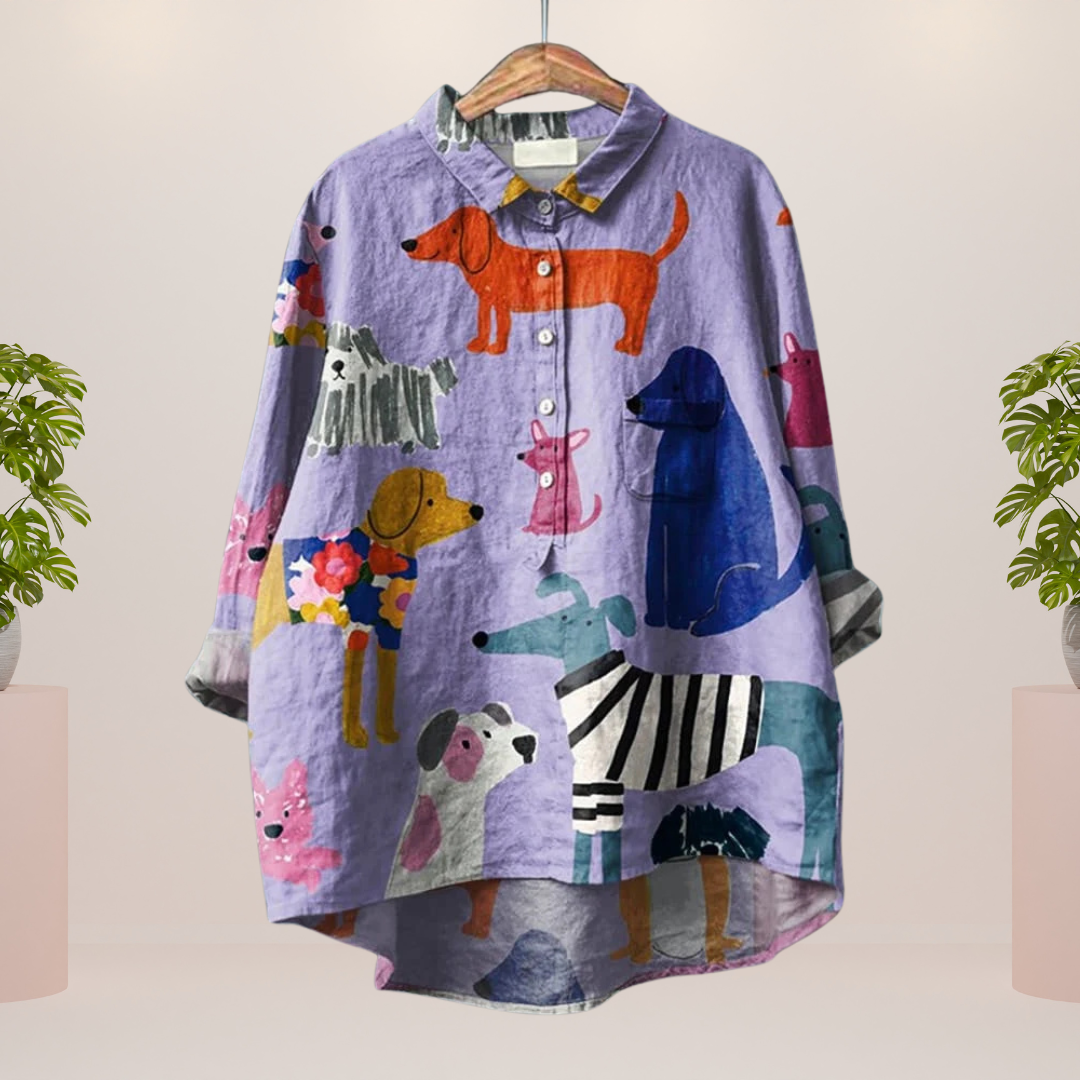 Camicia viola con stampa di cani colorati, design casual e moderno, appesa su gruccia. Sfondo neutro con piante decorative ai lati.
