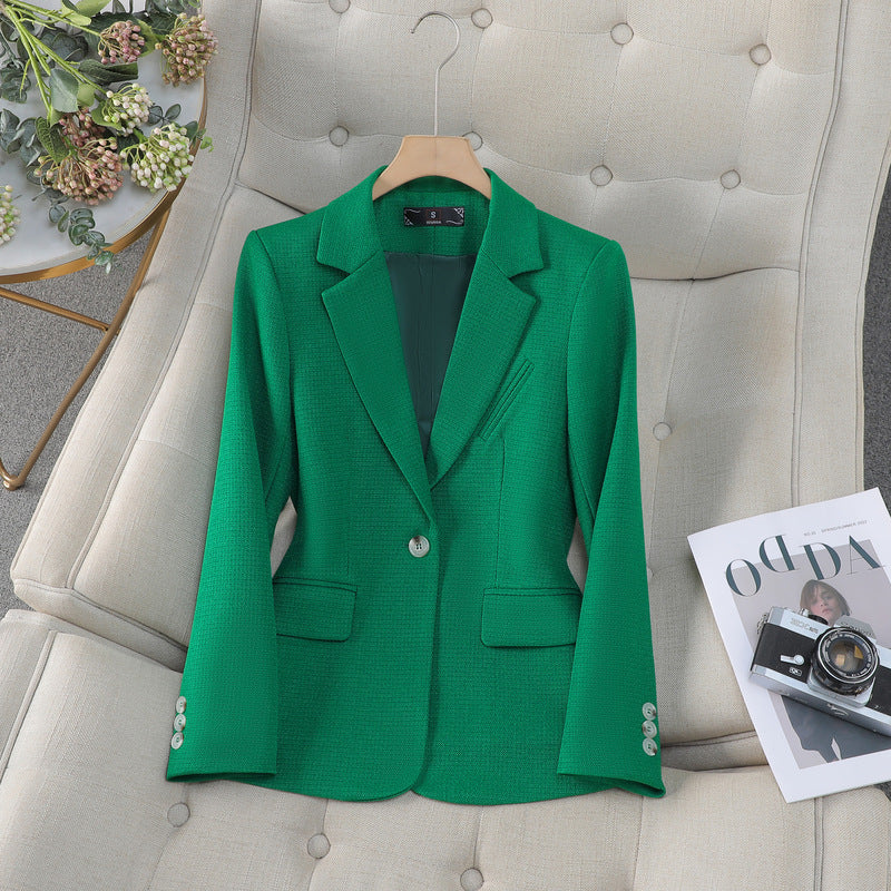 Giacca verde elegante su poltrona beige, accanto a rivista di moda e fiori decorativi. Abbigliamento formale, moda donna, stile sofisticato.