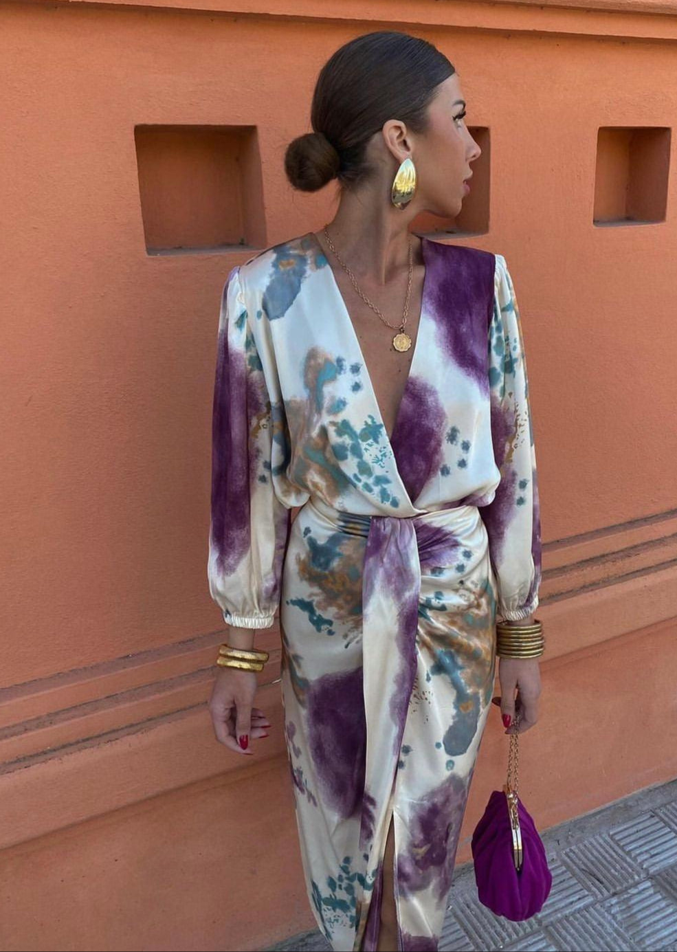 Donna con abito elegante a fantasia, orecchini dorati e borsa viola, posa davanti a muro arancione. Moda femminile, stile sofisticato.