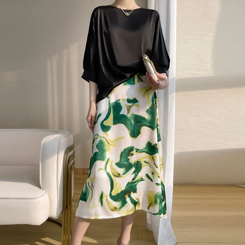 Donna in abito elegante con top nero e gonna a fantasia verde e gialla, in piedi accanto a una poltrona bianca, illuminata da luce naturale.