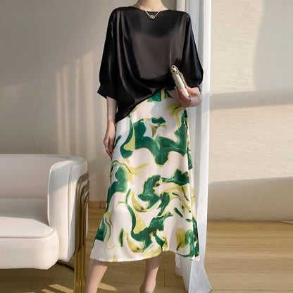 Donna in abito elegante con top nero e gonna a fantasia verde e gialla, in piedi accanto a una poltrona bianca, illuminata da luce naturale.