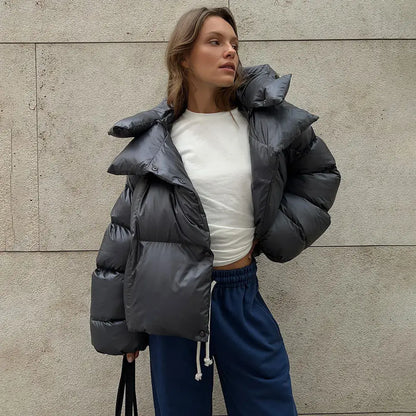 Donna con giacca piumino grigia oversize, maglia bianca e pantaloni sportivi blu, posa contro un muro. Moda invernale casual e trendy.