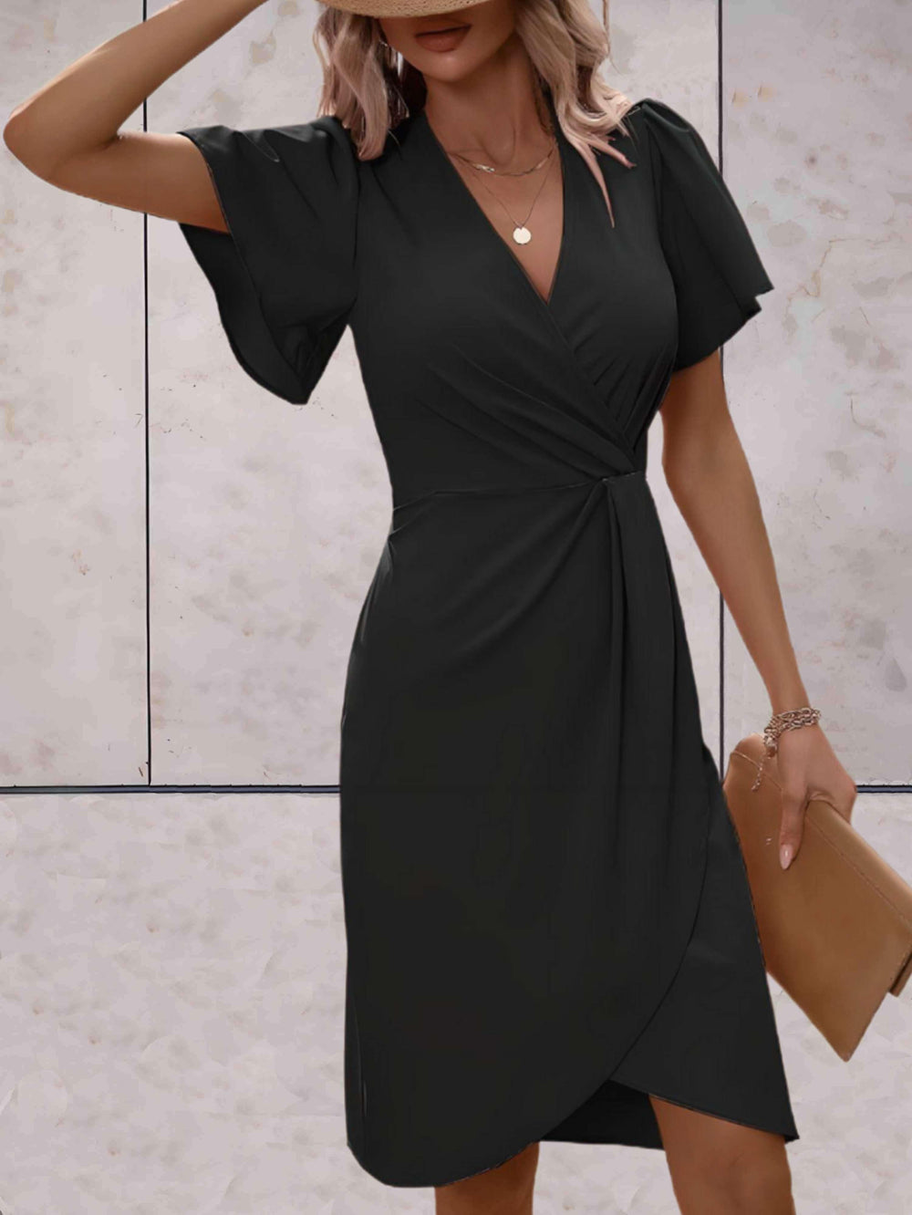Donna in abito nero elegante con scollo a V e maniche corte, tiene una pochette beige. Sfondo neutro. Moda femminile, abbigliamento chic.