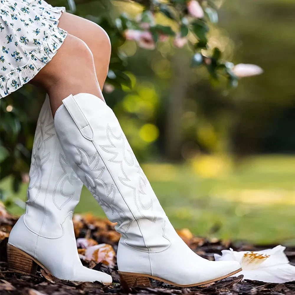 Stivali bianchi da cowboy in pelle con tacco basso, indossati da una donna in abito floreale, in un giardino verde. Moda autunnale, calzature eleganti.