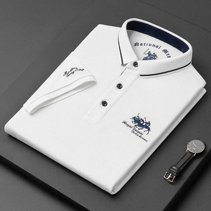 Camicia polo bianca piegata con colletto blu, logo ricamato, orologio nero e cinturino in pelle su sfondo nero. Elegante abbigliamento maschile.