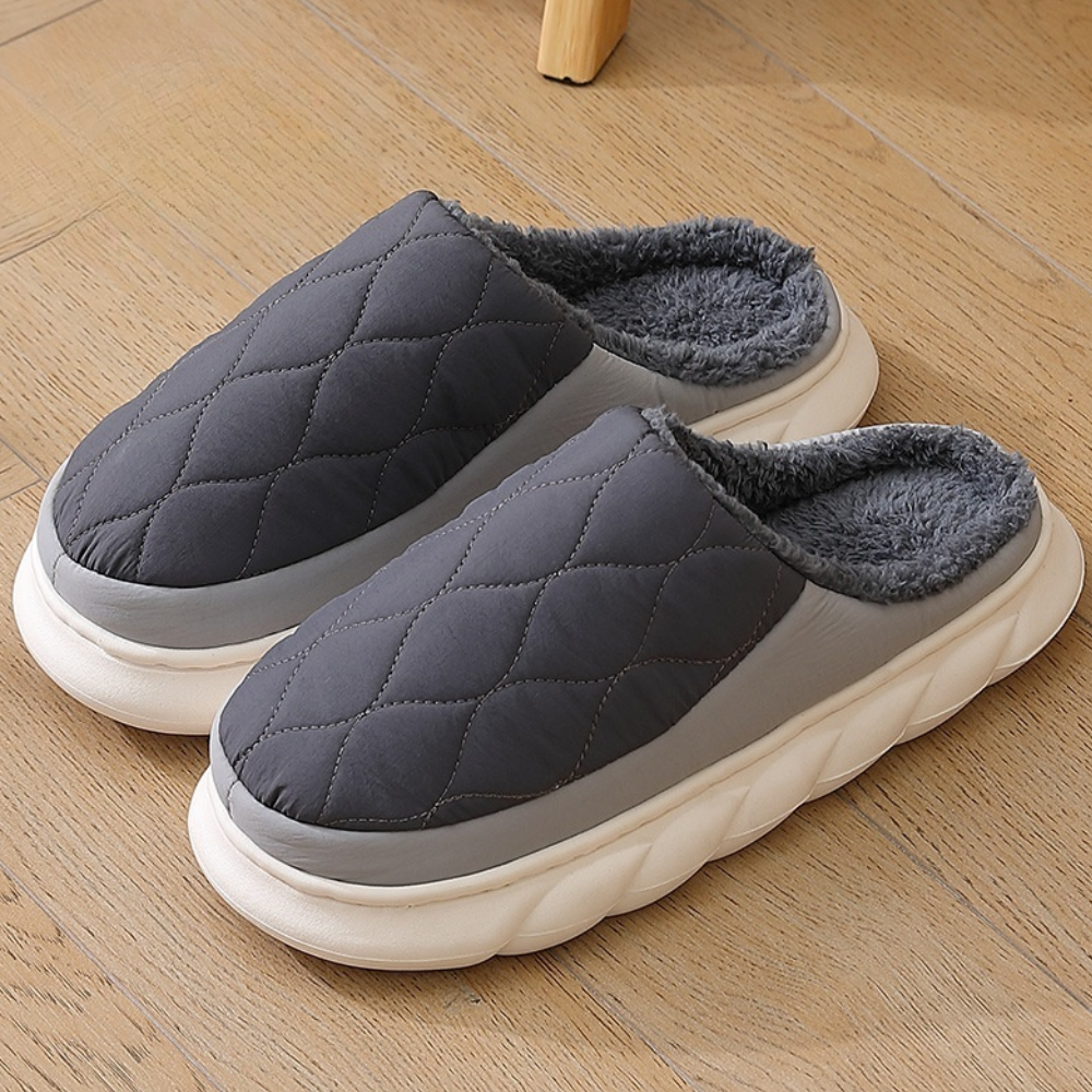 Pantofole imbottite grigie su pavimento in legno, design trapuntato, suola bianca spessa, interno in pelliccia sintetica, comfort invernale.