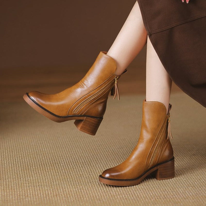 Stivali in pelle marrone con tacco medio, design elegante, chiusura con cerniera, indossati con abito marrone su sfondo neutro.