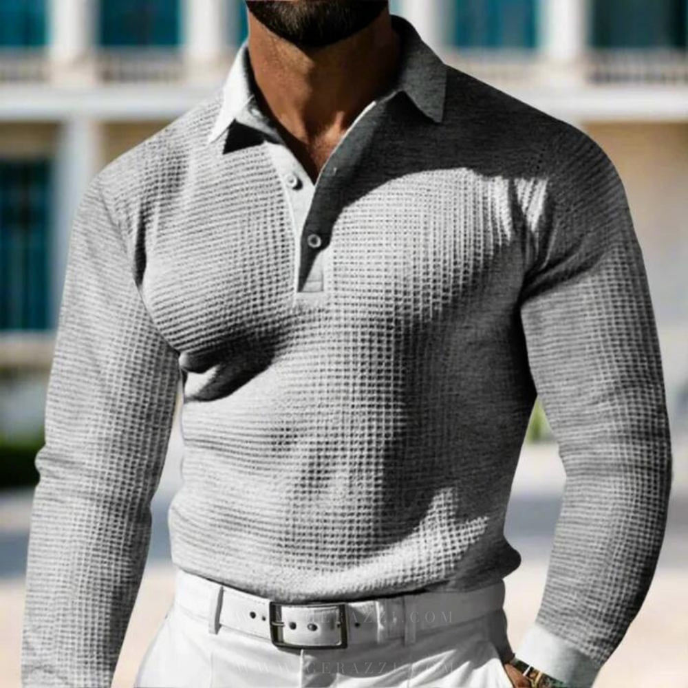 Uomo in camicia grigia a maniche lunghe con motivo a quadri, colletto e bottoni, indossa pantaloni bianchi. Sfondo urbano sfocato.