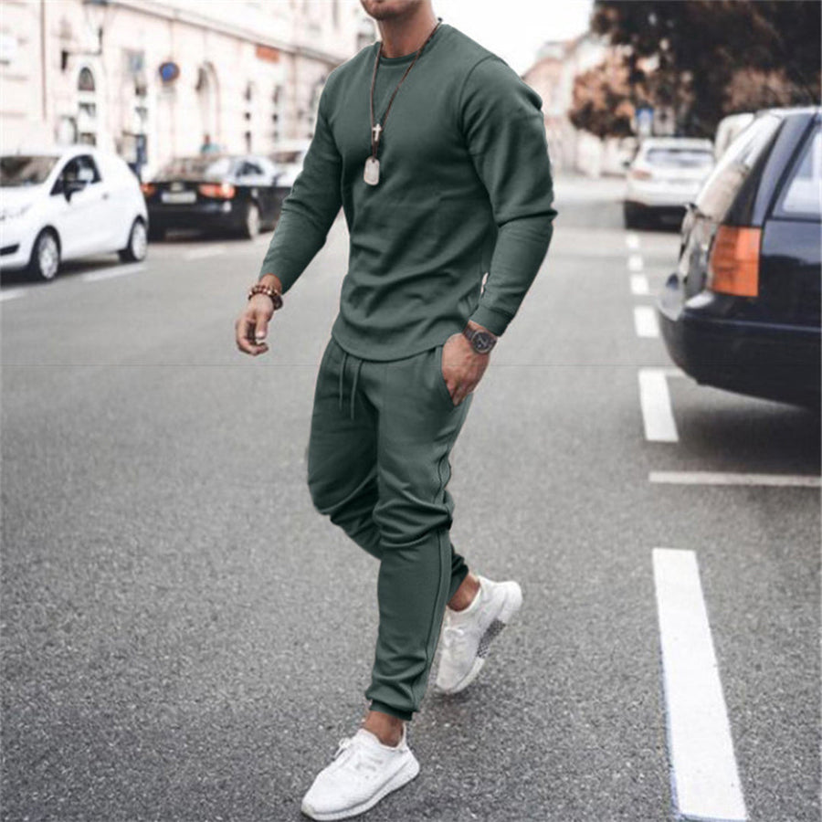 Uomo in tuta sportiva verde oliva, cammina su strada cittadina. Indossa scarpe da ginnastica bianche e orologio. Moda casual maschile urbana.
