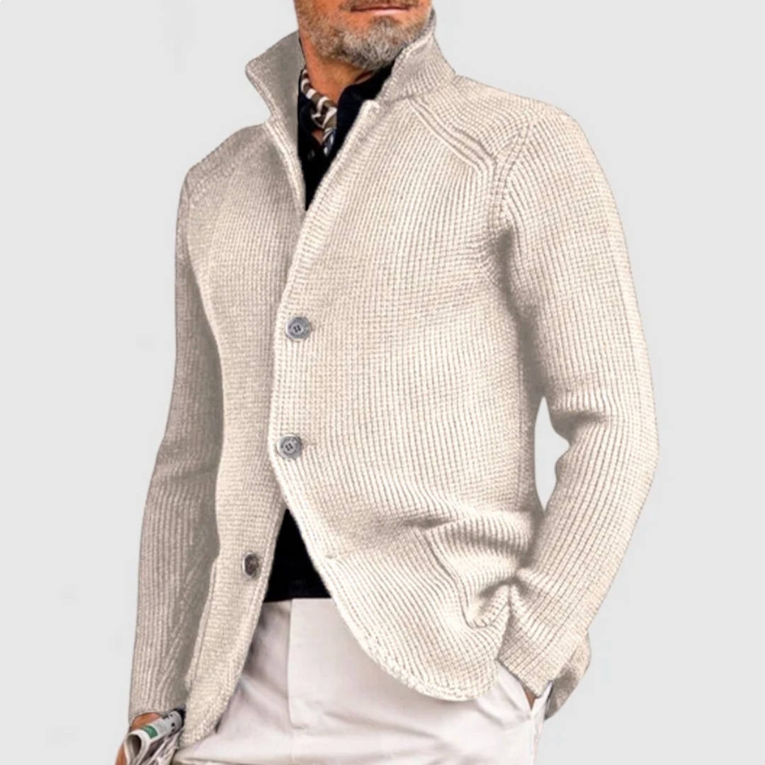 Uomo con cardigan beige a maglia grossa, colletto alto, abbinato a pantaloni chiari. Moda maschile elegante, stile autunnale.
