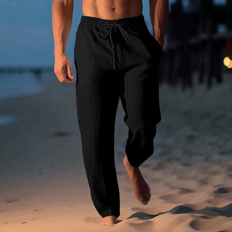 Uomo in pantaloni neri di lino cammina sulla spiaggia al tramonto. Abbigliamento estivo casual, moda maschile, relax in riva al mare.