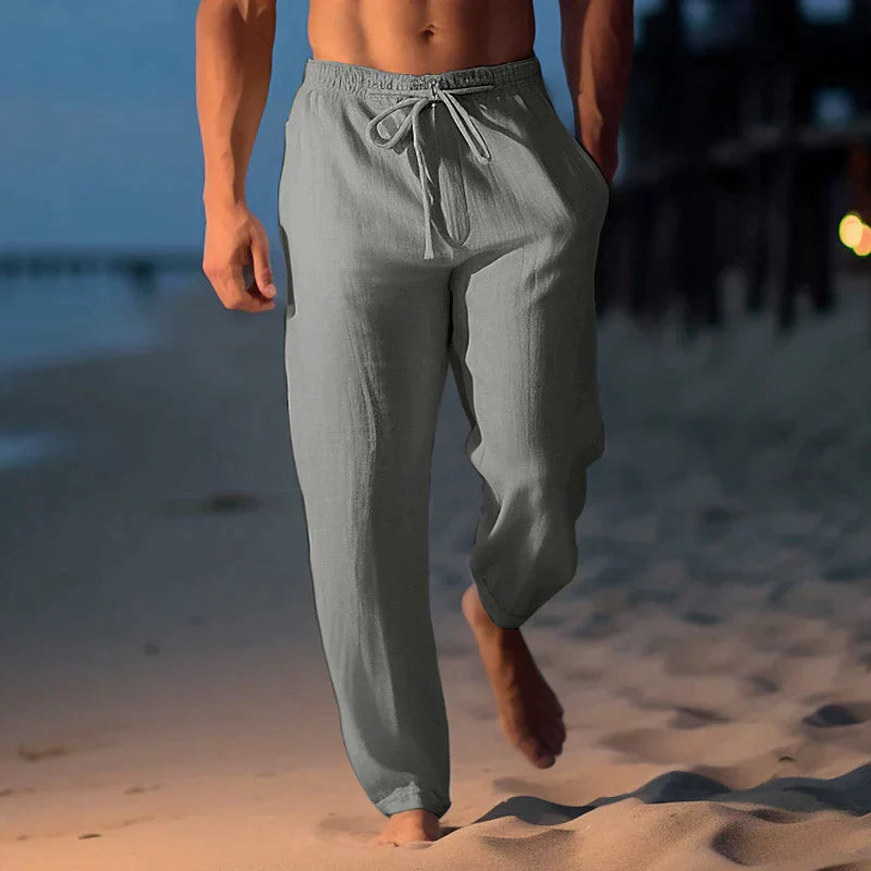 Uomo che cammina sulla spiaggia al tramonto indossando pantaloni di lino grigi, moda estiva maschile, abbigliamento casual elegante.