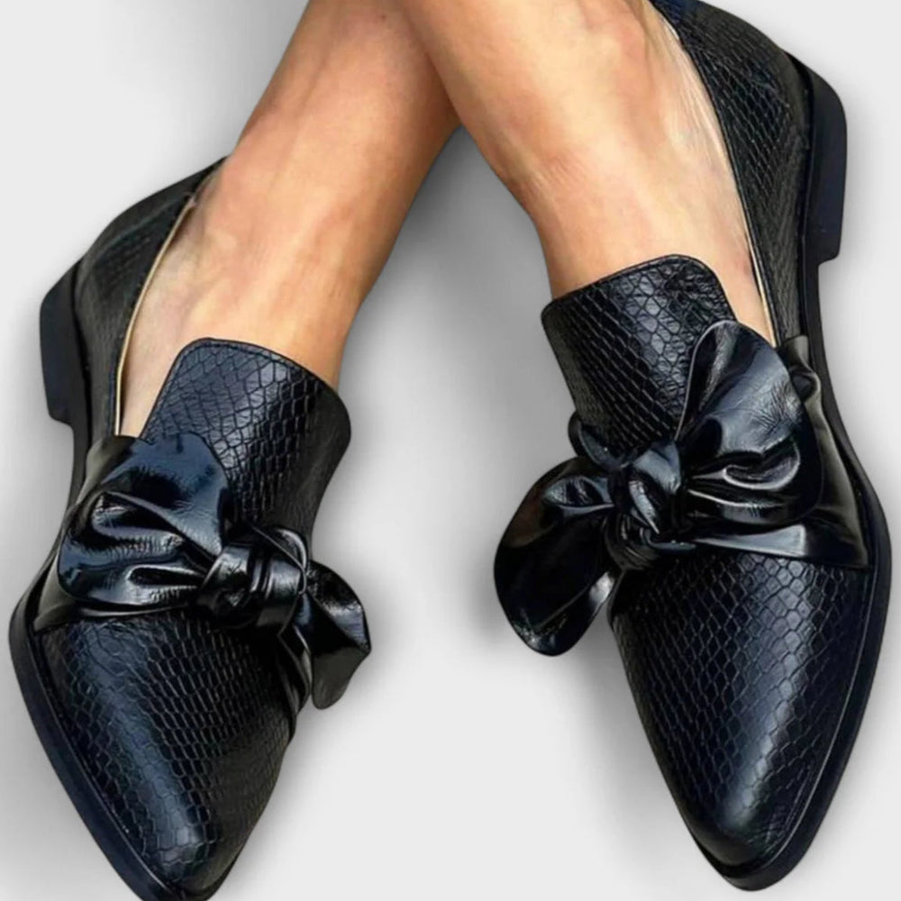 Scarpe eleganti nere in pelle con motivo a serpente e fiocco decorativo, indossate su gambe incrociate. Scarpe moda donna, stile sofisticato.