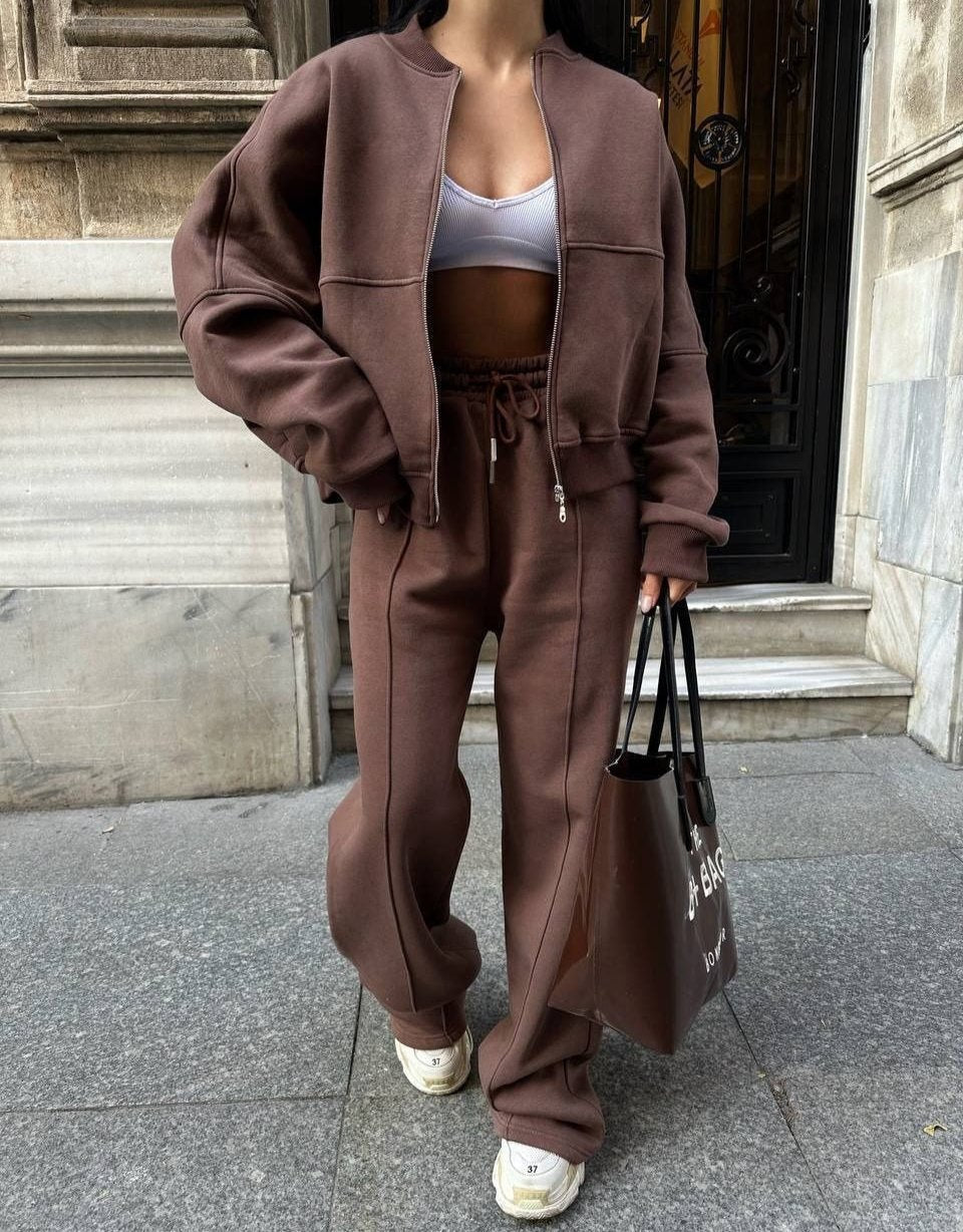 Donna con tuta marrone oversize, reggiseno sportivo bianco, borsa nera, scarpe da ginnastica bianche, davanti a un edificio storico.