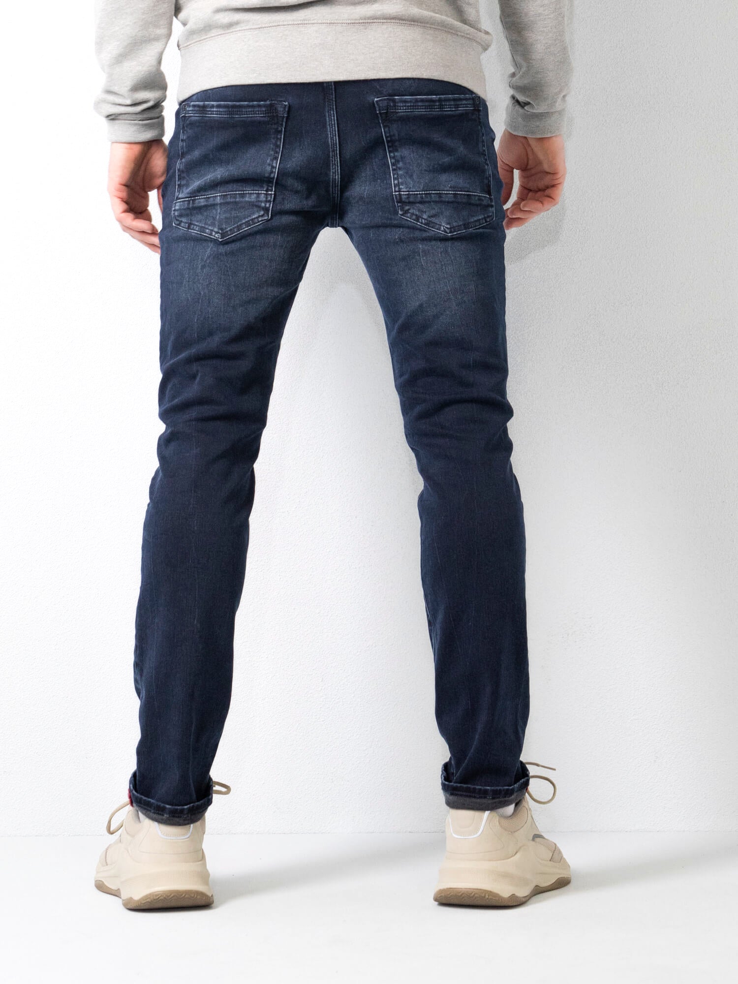 Jeans uomo slim fit blu scuro, vista posteriore, con tasche posteriori, abbinati a scarpe sportive beige, su sfondo bianco.