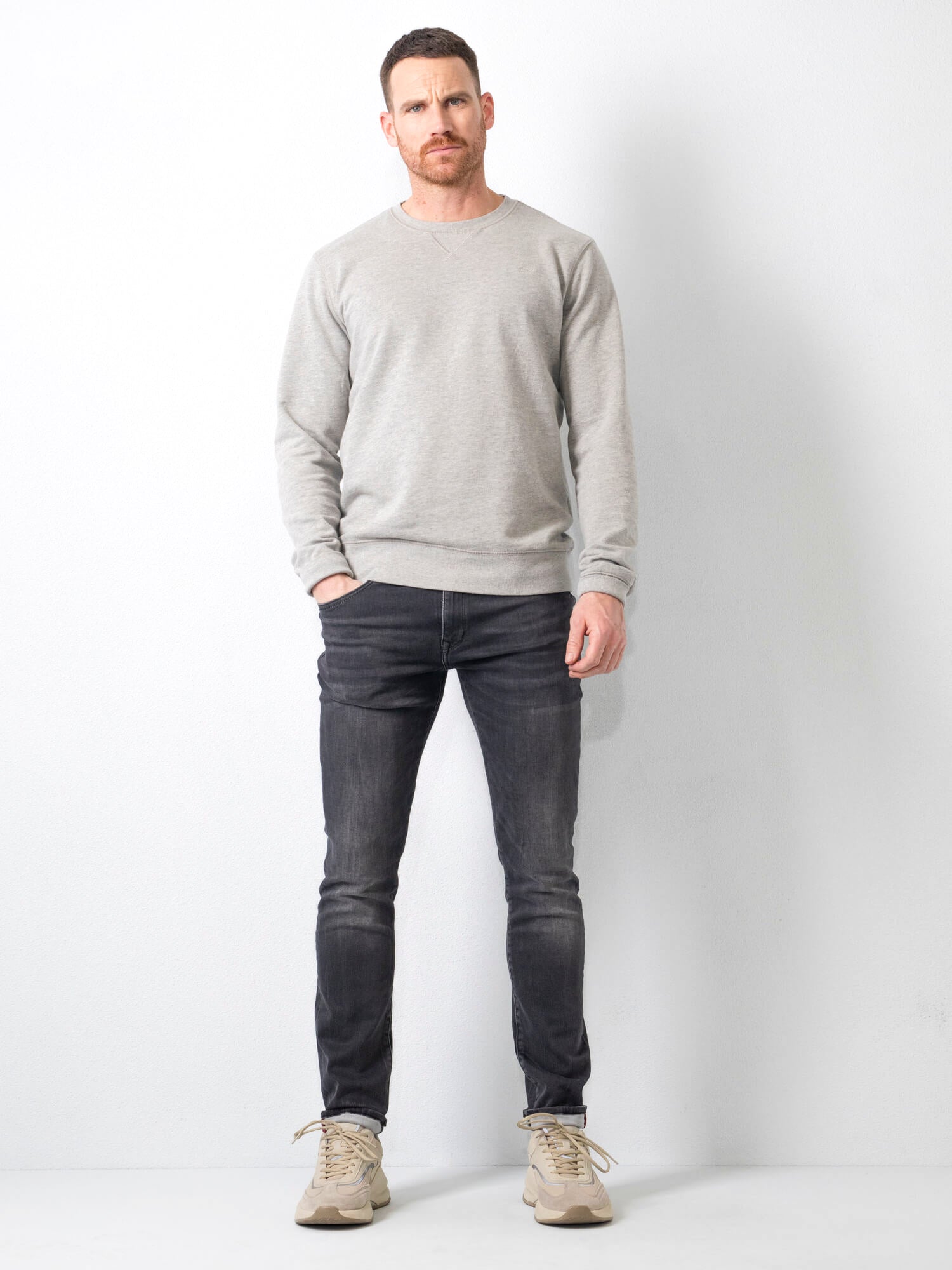 Uomo in posa con maglione grigio e jeans scuri, scarpe beige, sfondo bianco. Moda casual maschile, abbigliamento autunnale, stile moderno.