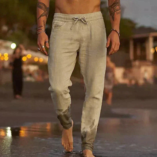 Uomo a torso nudo cammina sulla spiaggia al tramonto indossando pantaloni jogger beige, moda casual estiva, relax in riva al mare.
