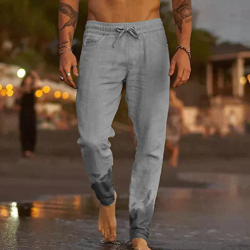 Uomo in pantaloni sportivi grigi cammina sulla spiaggia al tramonto, piedi bagnati. Abbigliamento casual estivo, moda maschile, relax in riva al mare.