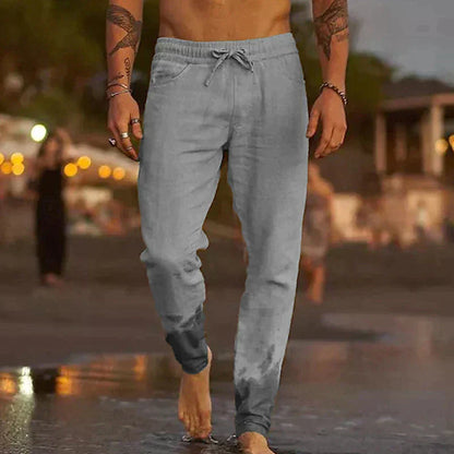 Uomo in pantaloni sportivi grigi cammina sulla spiaggia al tramonto, piedi bagnati. Abbigliamento casual estivo, moda maschile, relax in riva al mare.