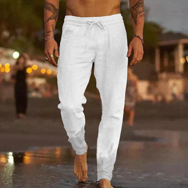Uomo che cammina sulla spiaggia al tramonto indossando pantaloni bianchi casual. Sfondo sfocato con luci e persone. Moda estiva maschile.