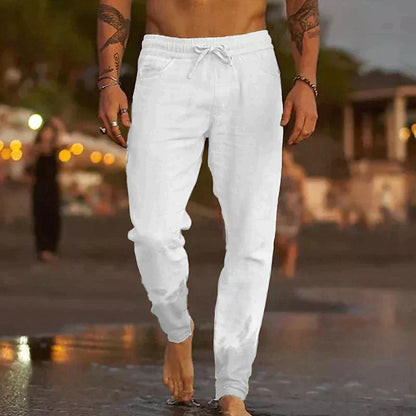 Uomo che cammina sulla spiaggia al tramonto indossando pantaloni bianchi casual. Sfondo sfocato con luci e persone. Moda estiva maschile.