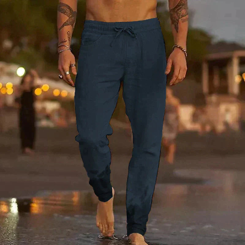 Uomo in pantaloni jogger blu scuro cammina a piedi nudi sulla spiaggia al tramonto, sfondo sfocato con luci e persone. Moda casual estiva.