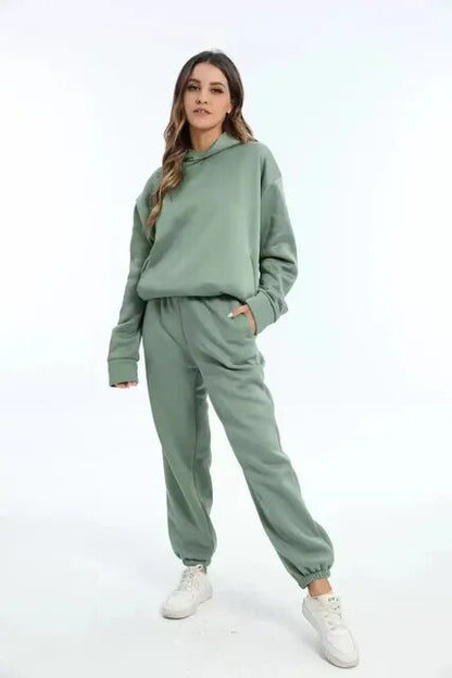 Donna in tuta sportiva verde salvia, felpa con cappuccio e pantaloni, scarpe da ginnastica bianche. Moda casual, abbigliamento comodo.