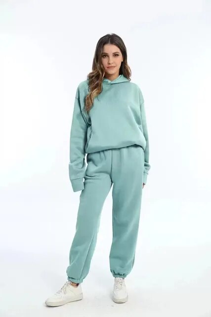 Donna in tuta sportiva verde acqua, felpa con cappuccio e pantaloni abbinati, scarpe da ginnastica bianche, moda casual, sfondo bianco.