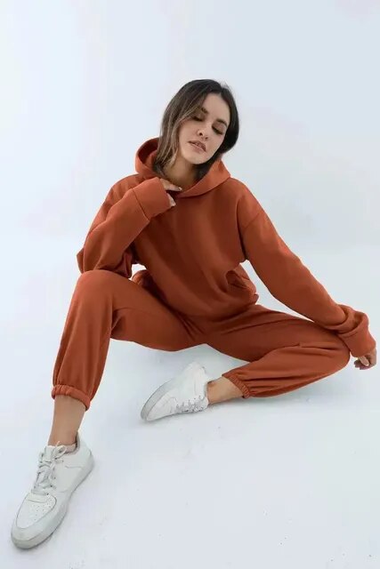 Donna in tuta sportiva marrone, seduta su sfondo bianco. Indossa felpa con cappuccio e pantaloni abbinati, scarpe da ginnastica bianche. Moda casual.