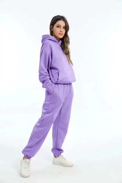 Donna in tuta sportiva viola, felpa con cappuccio e pantaloni abbinati, posa su sfondo bianco. Moda casual, abbigliamento comodo.