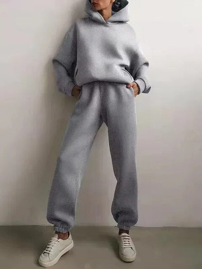 Donna in tuta grigia oversize con cappuccio, pantaloni larghi e sneakers bianche. Moda casual, abbigliamento comodo, stile urbano.