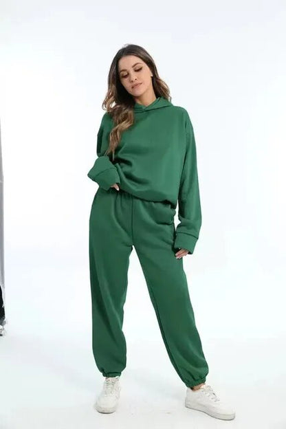 Donna in tuta verde, felpa con cappuccio e pantaloni abbinati, posa su sfondo bianco. Moda casual, abbigliamento comodo, stile sportivo.