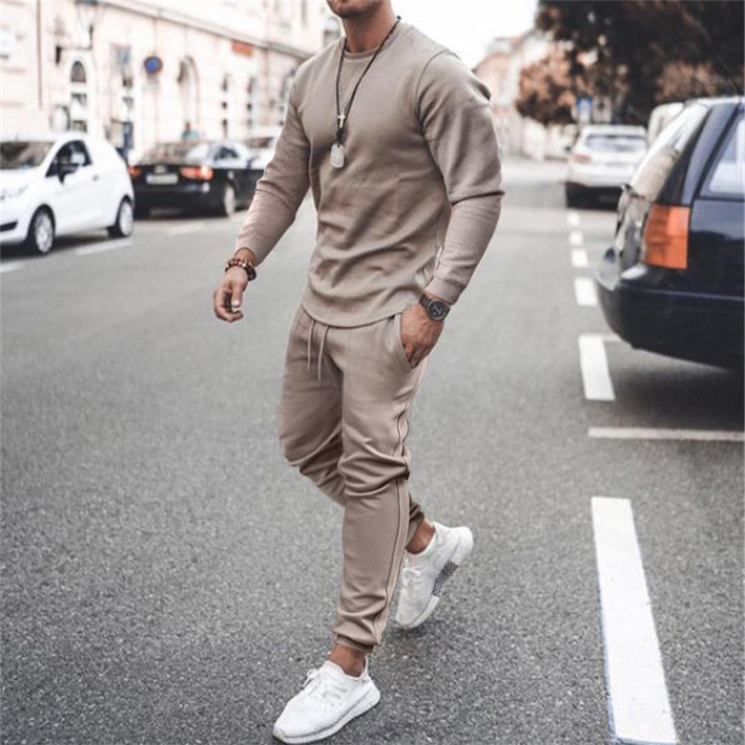 Uomo in tuta sportiva beige cammina su strada cittadina, indossa scarpe da ginnastica bianche, orologio e collana. Moda casual urbana.