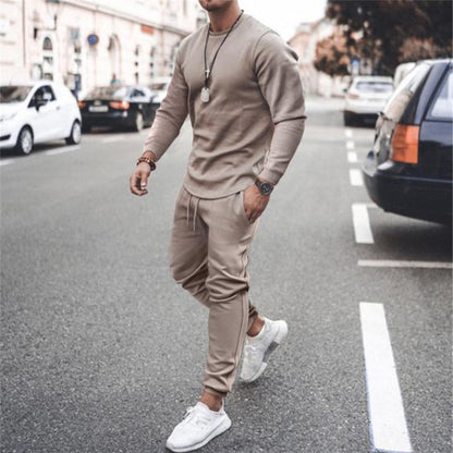 Uomo in tuta sportiva beige cammina su strada cittadina, indossa scarpe da ginnastica bianche, orologio e collana. Moda casual urbana.