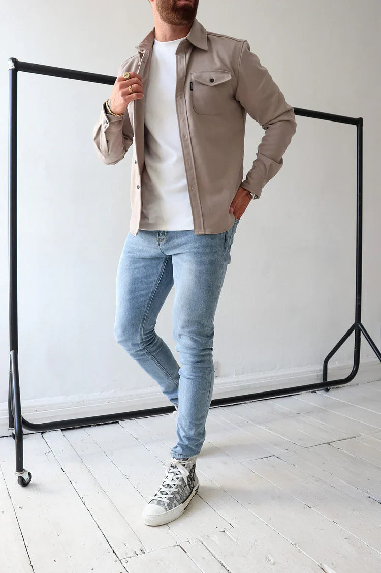 Uomo in jeans attillati e camicia beige, posa su pavimento in legno bianco. Moda casual maschile, scarpe da ginnastica con stampa.
