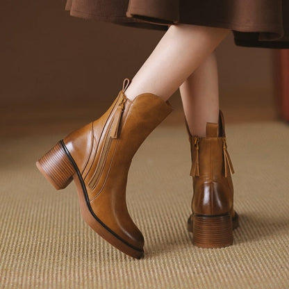 Stivali in pelle marrone con tacco largo, chiusura a zip laterale e dettagli con frange, su pavimento beige. Moda autunno, calzature eleganti.