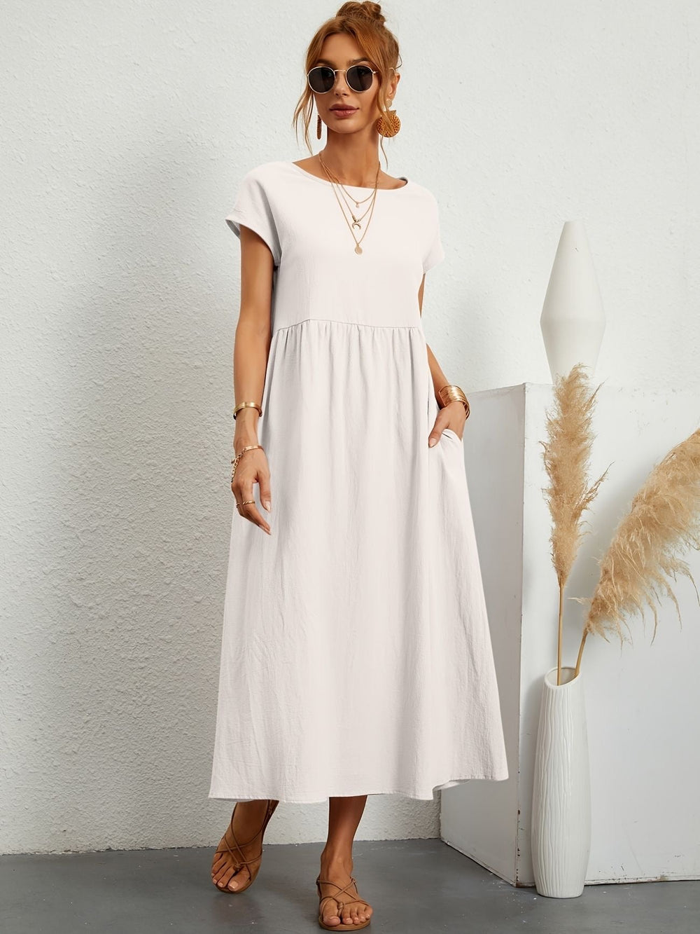 Donna con abito lungo bianco, occhiali da sole e sandali, accanto a vaso decorativo con pampas. Moda estiva elegante e casual.