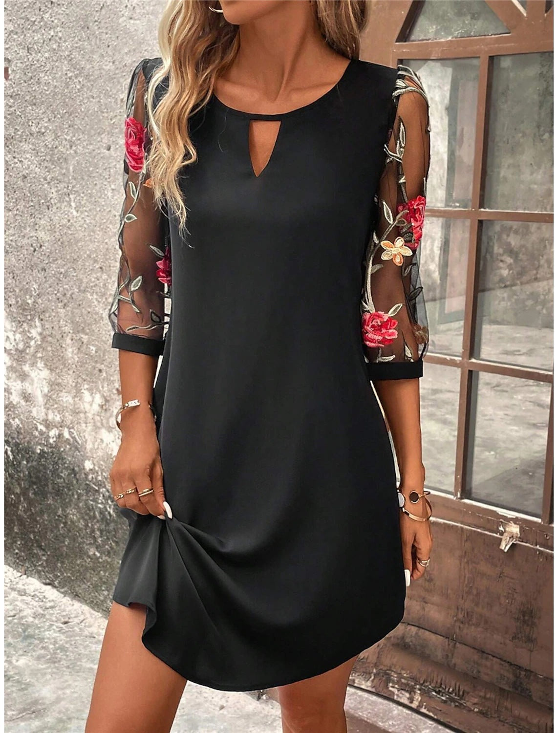 Donna con abito nero elegante, maniche trasparenti ricamate con fiori rossi e bianchi, posa vicino a una finestra. Moda femminile, abbigliamento chic.