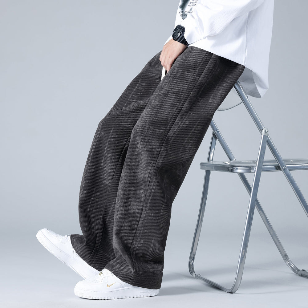 Pantaloni larghi in velluto a coste neri, indossati con scarpe da ginnastica bianche e una camicia bianca, seduto su una sedia pieghevole.