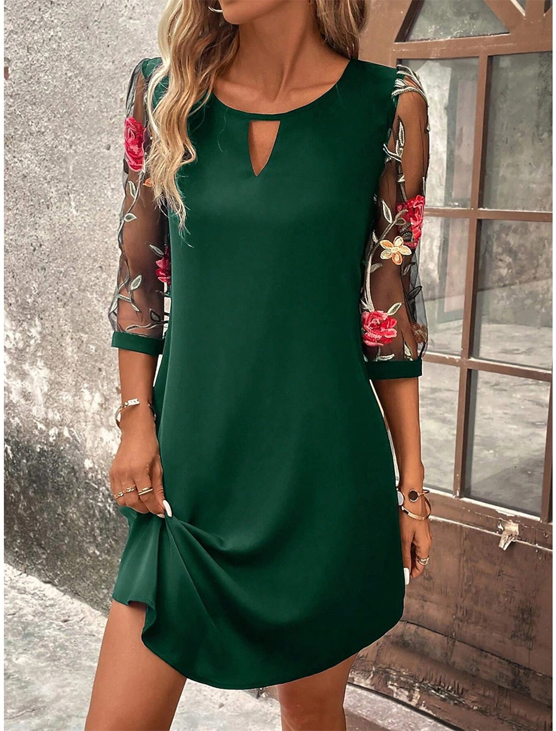 Donna con abito verde elegante, maniche trasparenti ricamate con fiori rossi e bianchi, posa vicino a una finestra. Moda femminile, abbigliamento chic.