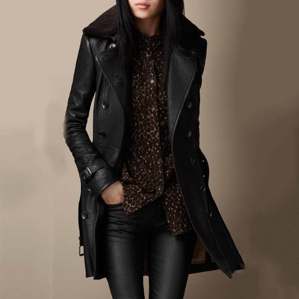 Giacca in pelle nera da donna, stile trench, con bottoni e colletto in pelliccia, abbinata a camicia marrone e pantaloni neri. Moda autunnale elegante.
