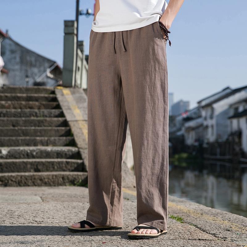 Pantaloni larghi in lino marrone, indossati con sandali neri, su strada acciottolata vicino a canale. Moda estiva casual uomo, comfort e stile.