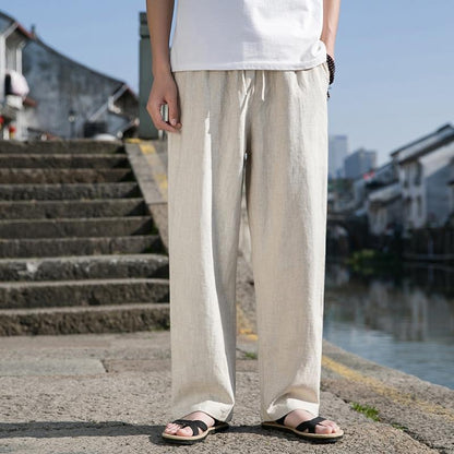 Uomo in pantaloni di lino beige e sandali neri su strada acciottolata vicino a canale, sfondo urbano. Moda estiva casual, comfort, stile rilassato.