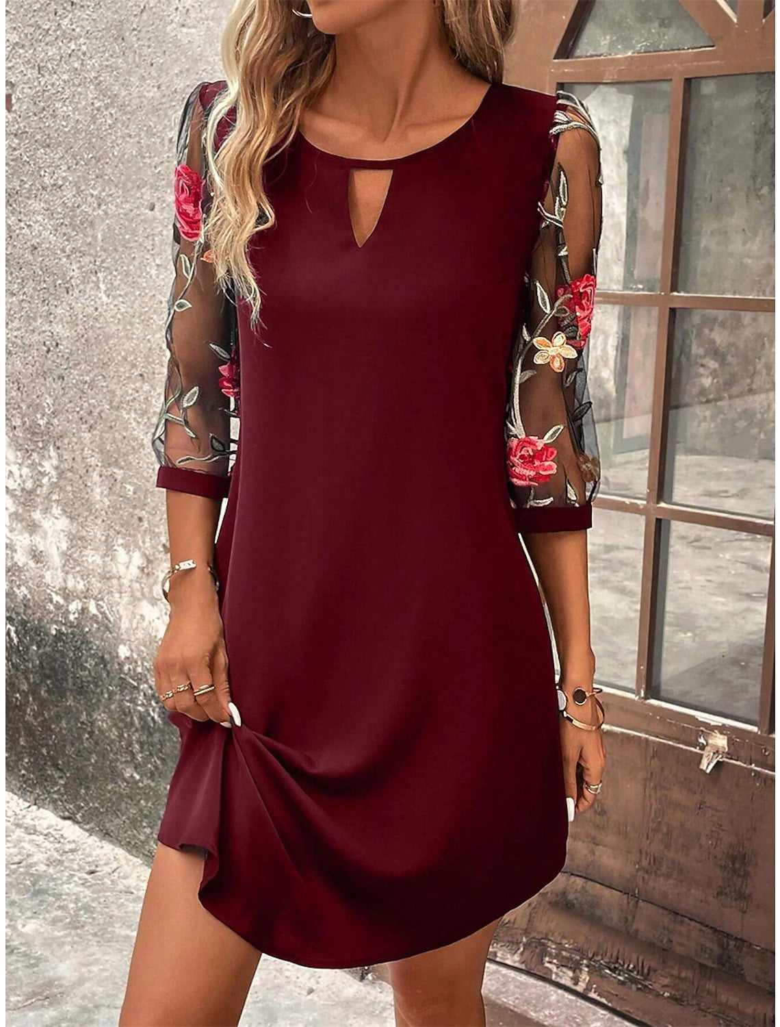 Donna con abito bordeaux elegante, maniche trasparenti ricamate con fiori, posa vicino a una finestra. Moda femminile, abbigliamento autunnale chic.