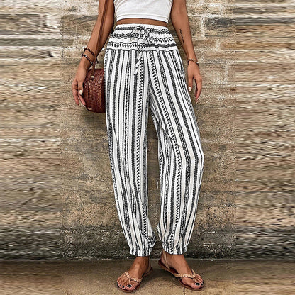 Pantaloni a righe bianche e nere, stile boho, con cintura annodata, indossati da una donna con top bianco e borsa intrecciata marrone.