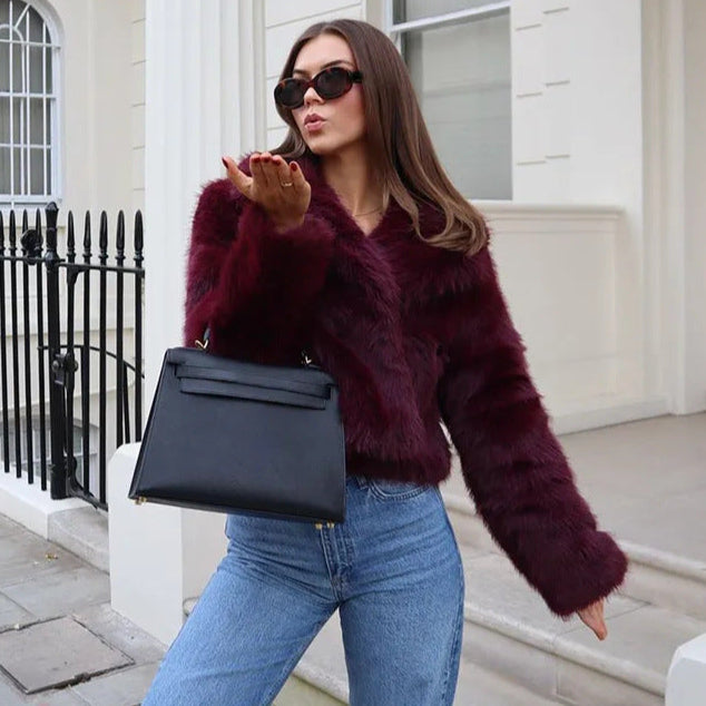 Donna con giacca in pelliccia bordeaux, occhiali da sole neri, jeans blu e borsa nera, posa all'aperto. Moda autunnale elegante e chic.
