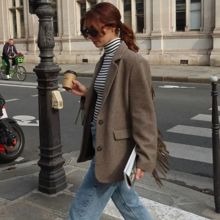 Donna con occhiali da sole, giacca marrone, maglia a righe e jeans, cammina su marciapiede con caffè in mano. Sfondo urbano con lampione.